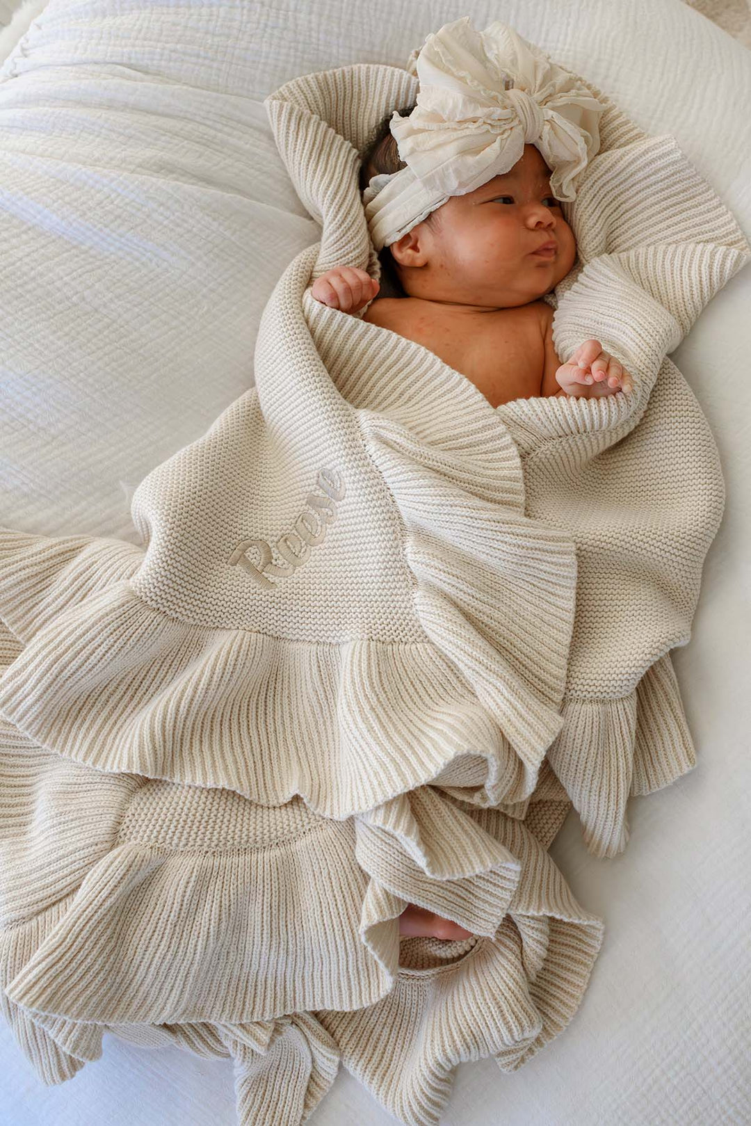 A newborn baby wrapped in a cream-colored ribbed knit blanket with ruffles, wearing a matching fabric head wrap, lying on a white textured blanket.