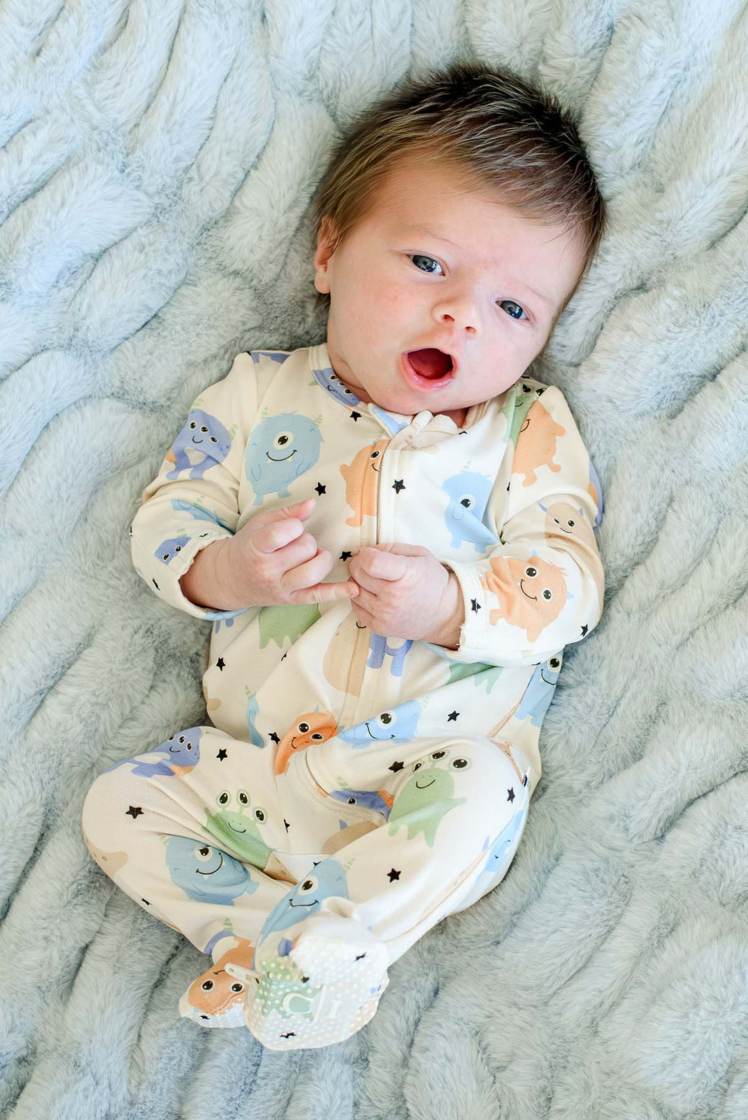 Newborn laying on furry blue blanket wearing a Caden Lane Little Monsters zipper footie in cream with blue, green, and orange cartoon monsters