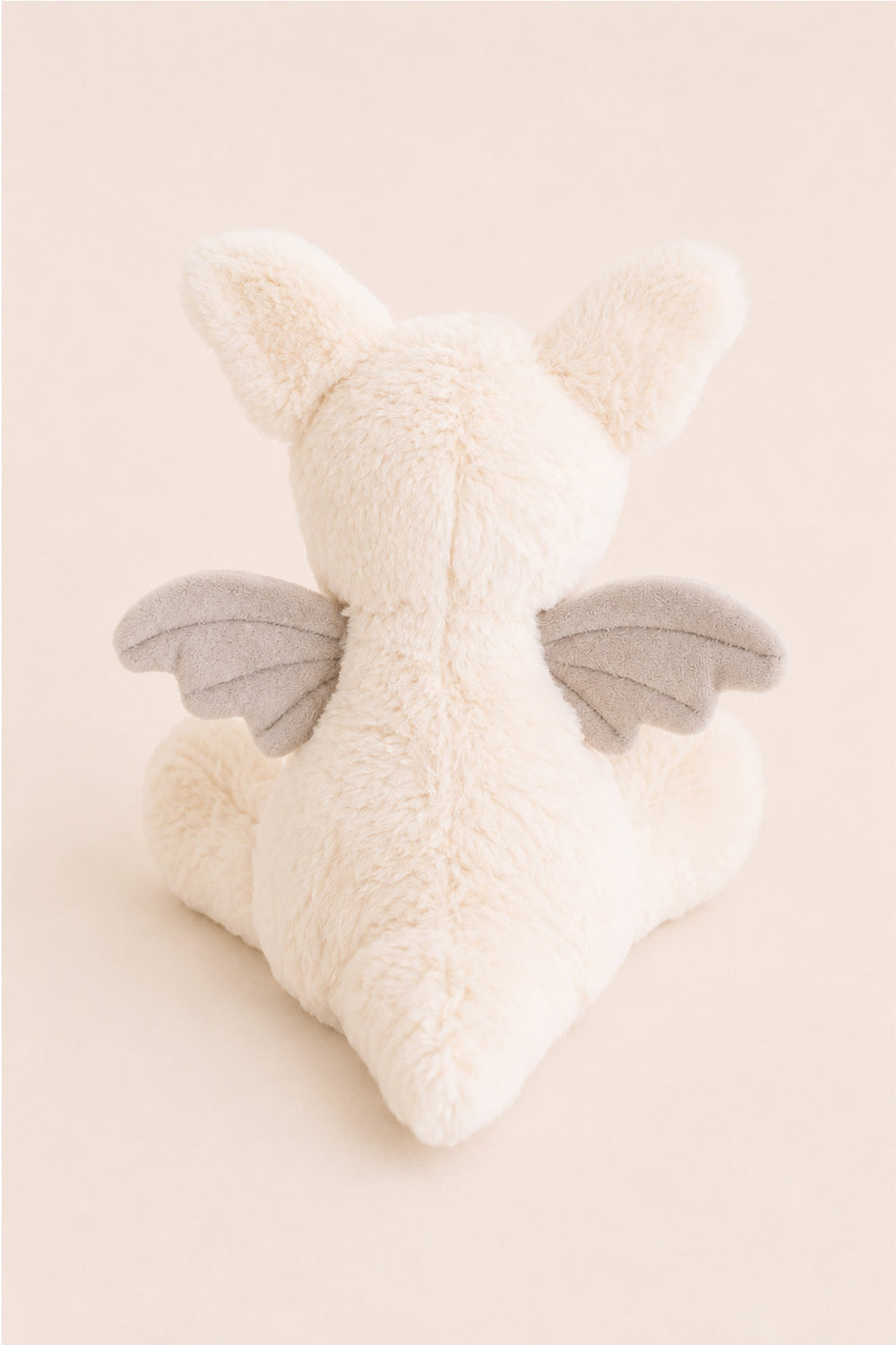 A Caden Lane plush toy dragon shown from the back, featuring fluffy cream-colored fur, small pointed ears, and soft gray wings, set against a solid pale beige background.