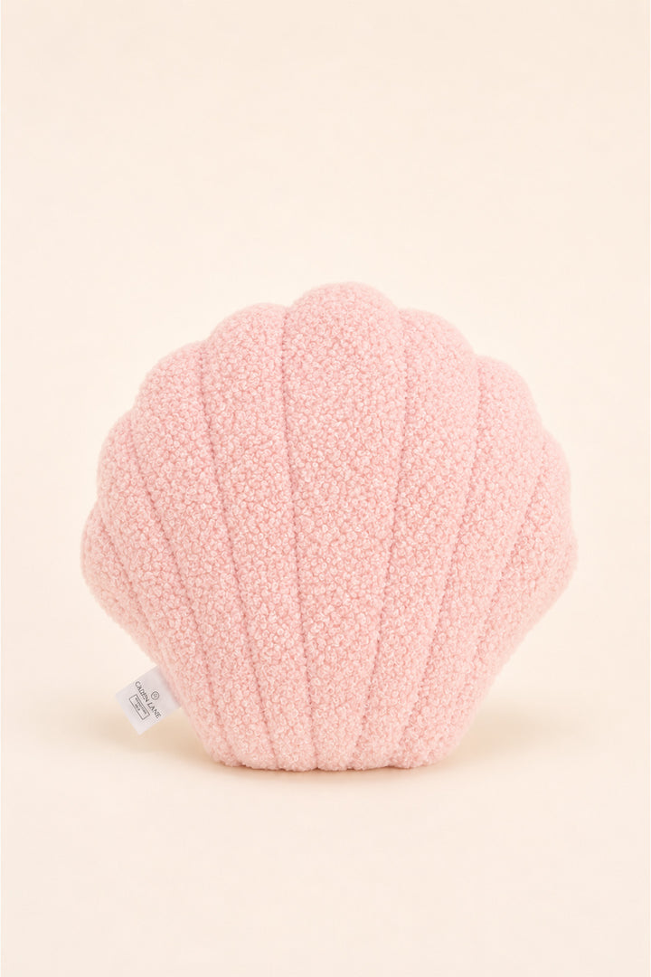 A Caden Lane light pink plush pillow shaped like a seashell, featuring a ribbed texture and a small white fabric tag on the lower left, set against a solid cream background.