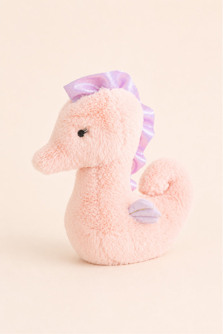 A Caden Lane plush seahorse toy in soft pink with a lavender-purple ribbon mane and a matching small tail fin, photographed on a plain warm cream background.