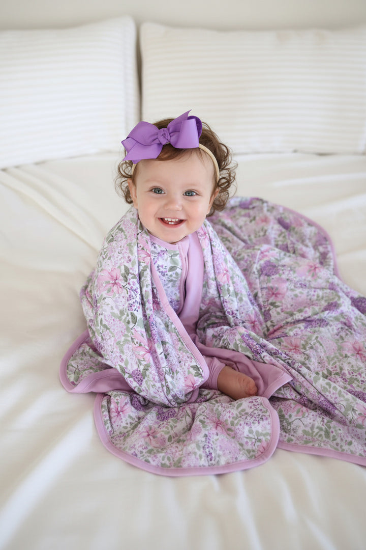 sitting baby girl with lavender lullaby bamboo blanket around her shoulders wearing matching pajamas and a purple ribbon bow headband