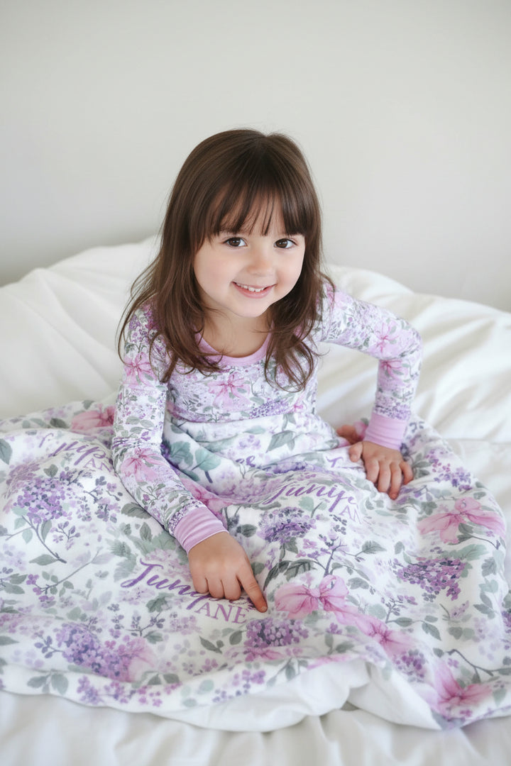 girl wearing lavender lullaby two piece pajama set with a matching personalized blanket across her lap 