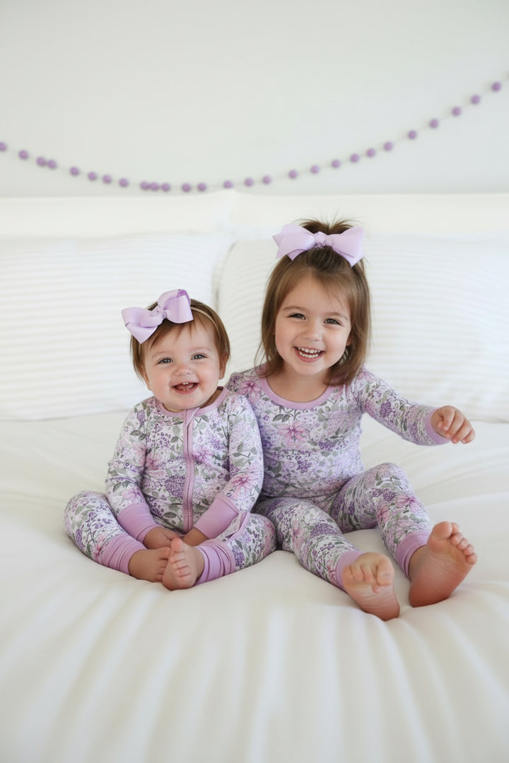 sisters sitting on bed in lavender lullaby convertible zip romper and two piece pajama set