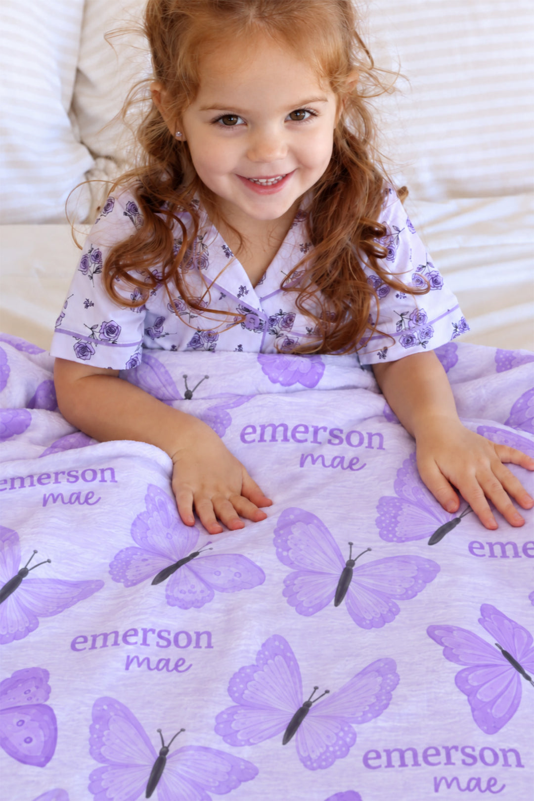 Smiling girl in floral pajamas sitting on a bed with hands resting on a personalized Caden Lane Lavender Wings blanket, showing purple butterflies and custom name print