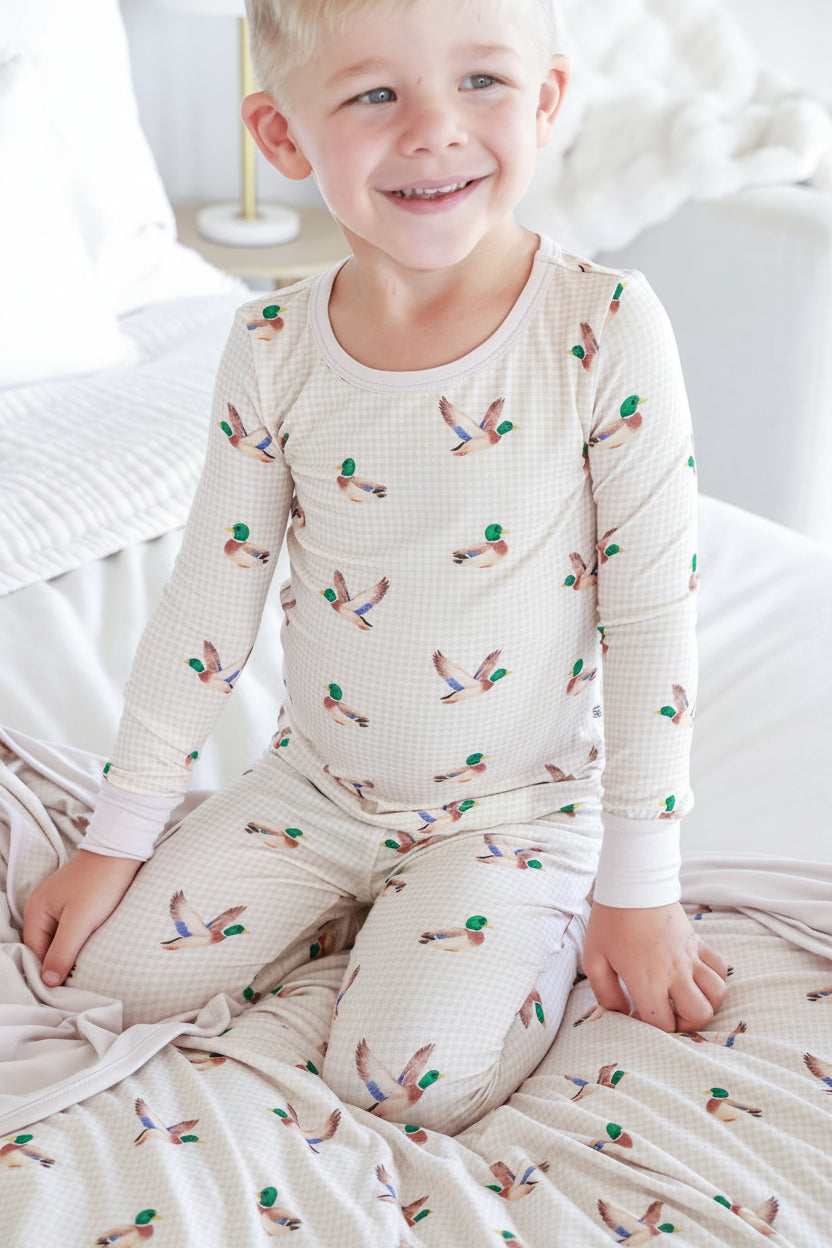 Child sitting on bed smiling wearing little mallard duck print two-piece bamboo pajama set with long-sleeve top and pants