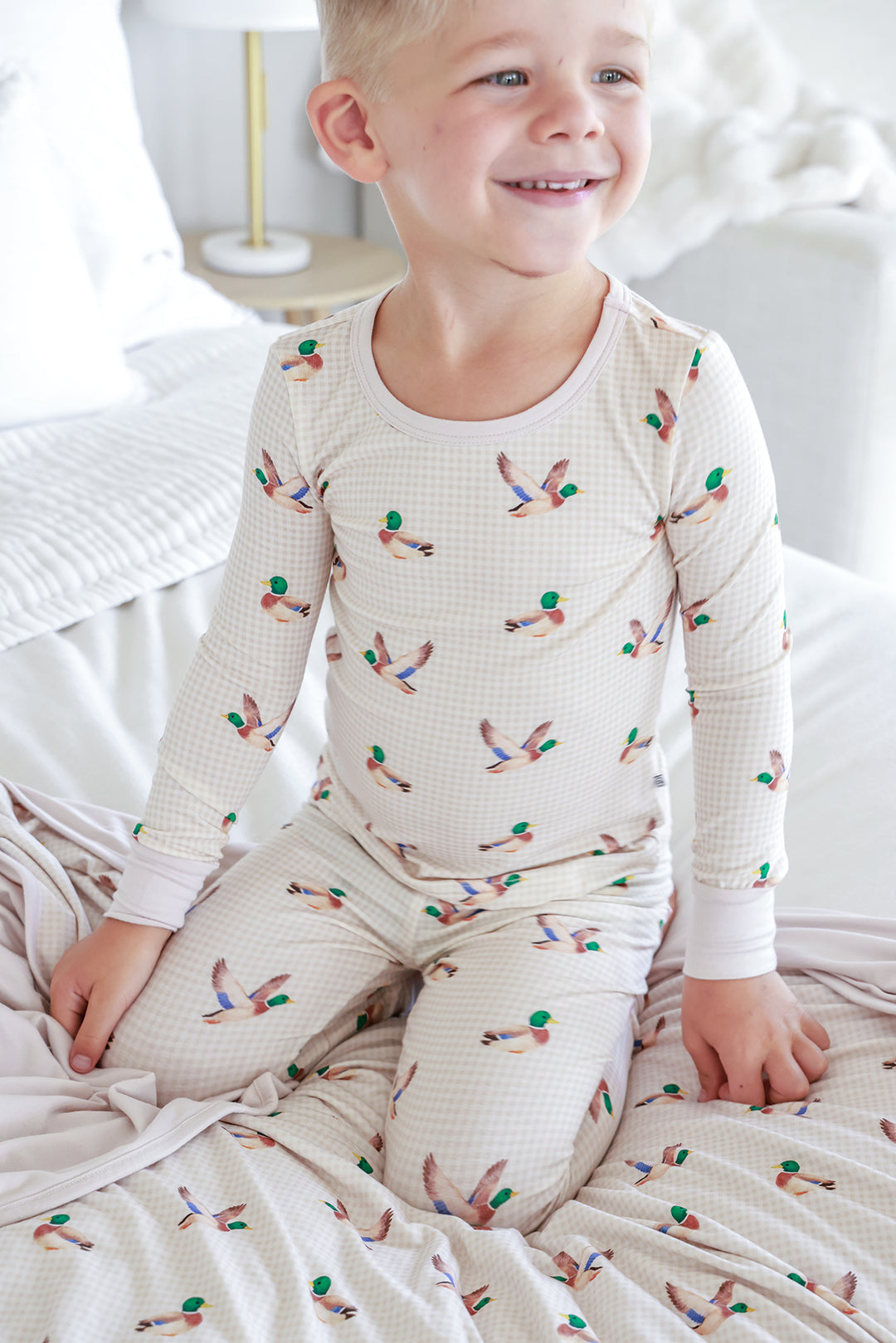 Smiling child sitting on bed wearing little mallard duck print two-piece bamboo pajama set, full view showing top and pants