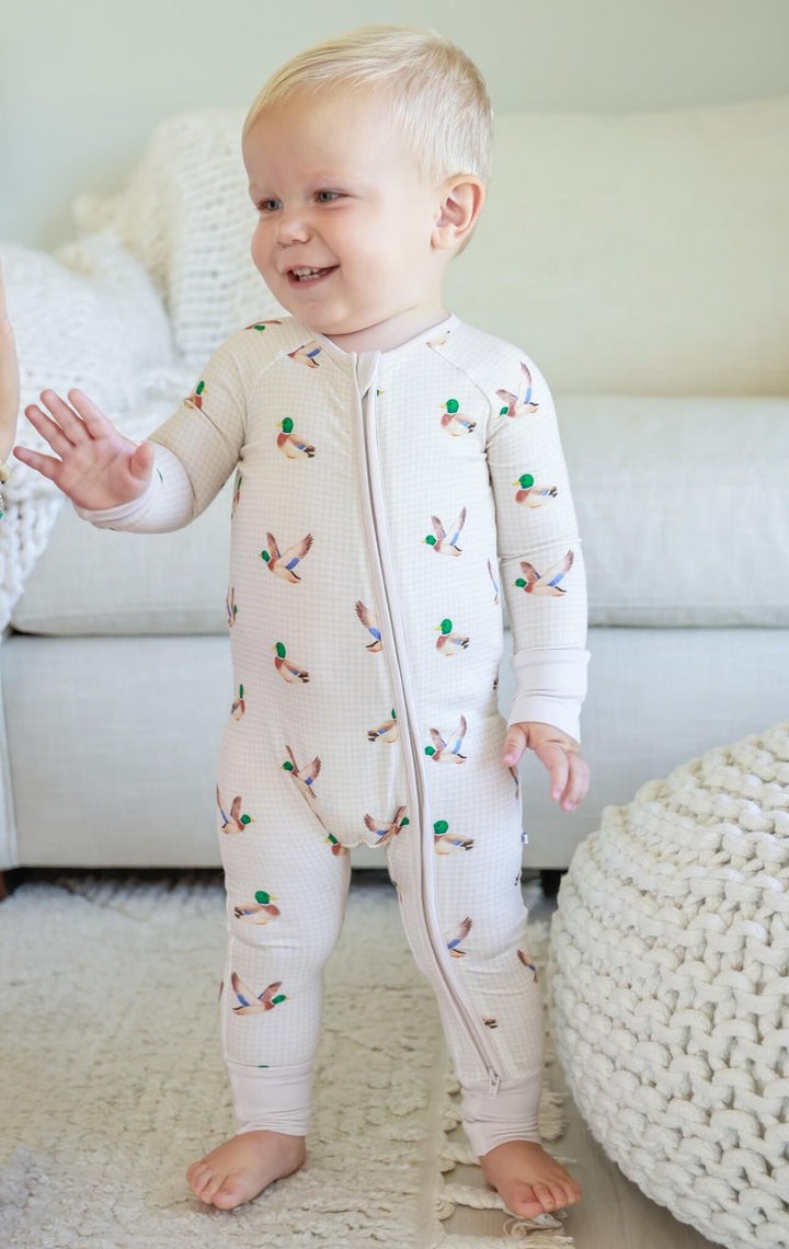 Toddler standing wearing little mallard duck print convertible zip romper, waving hand, showing full-length front view