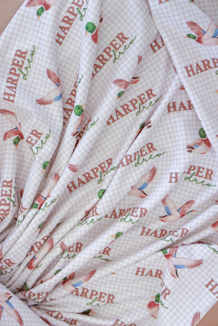 Close-up detail of personalized little mallard duck print swaddle blanket fabric showing name and flying duck pattern on gingham background