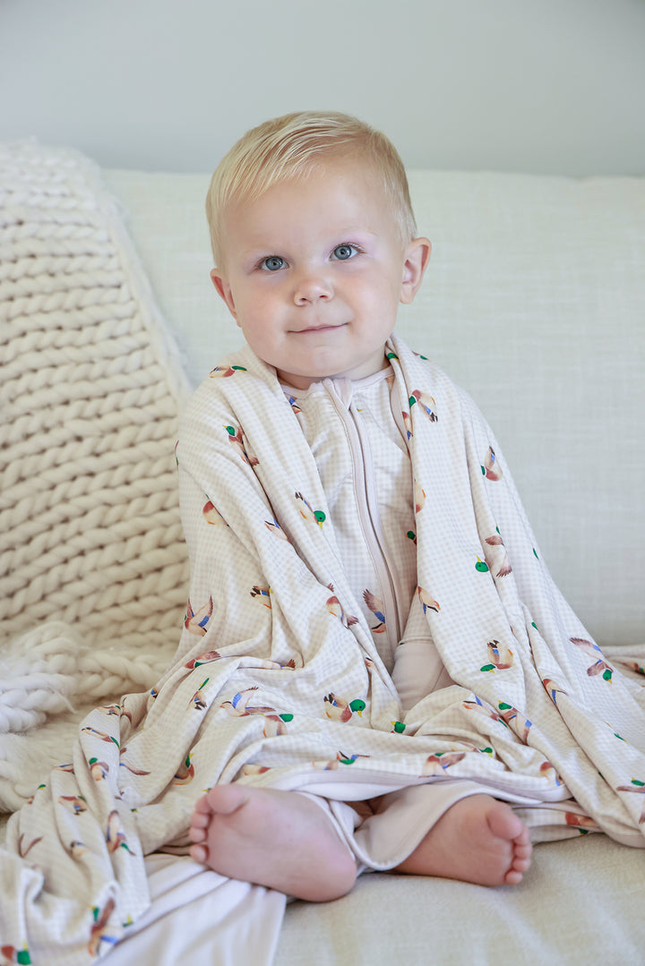 Smiling toddler lying on bed wrapped in little mallard duck print double-sided bamboo blanket, overhead view