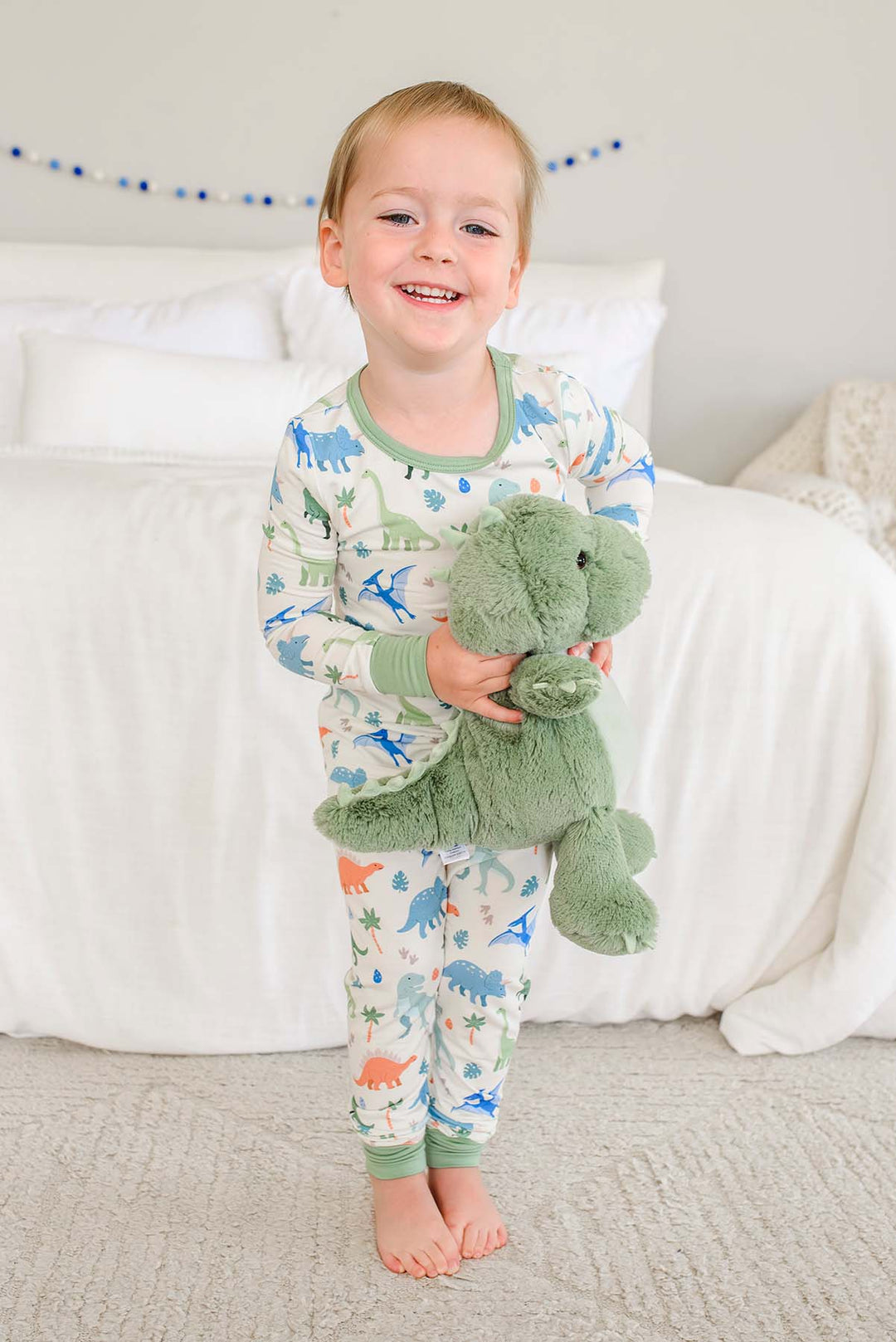 smiling boy standing in front of bed in little fossil neutral two piece pajama set holding a large stuffed animal