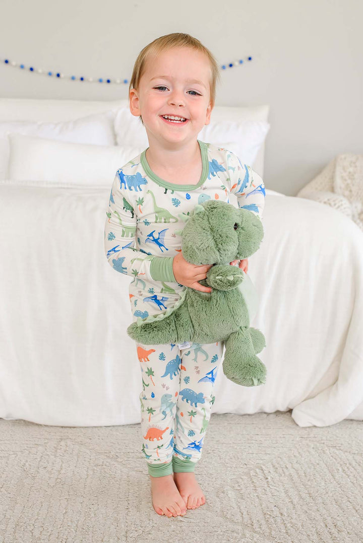 smiling boy standing in front of bed in little fossil neutral two piece pajama set holding a large stuffed animal