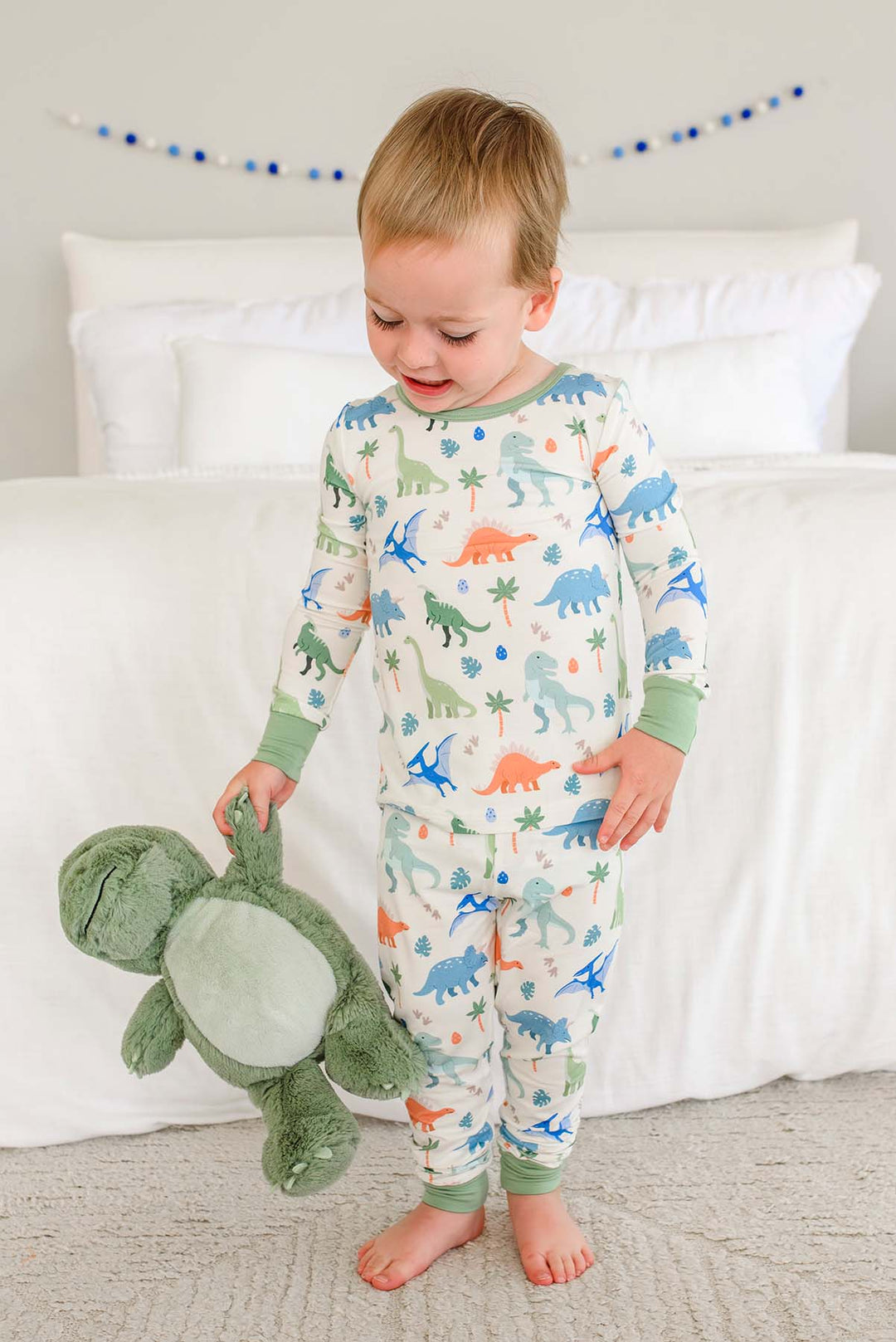 standing boy smiling wearing dinosaur pajamas and holding a stuffed t-rex