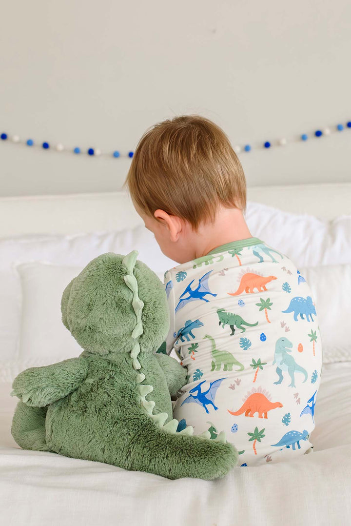 boy sitting on bed in dinosaur pajamas next to dinosaur stuffed animal 
