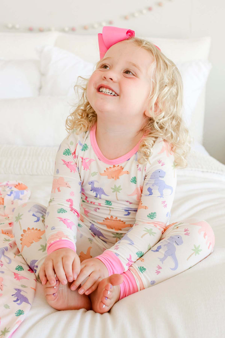 girl sitting on bed smiling wearing little fossil pink two piece pajama set with a pink ribbon clip bow