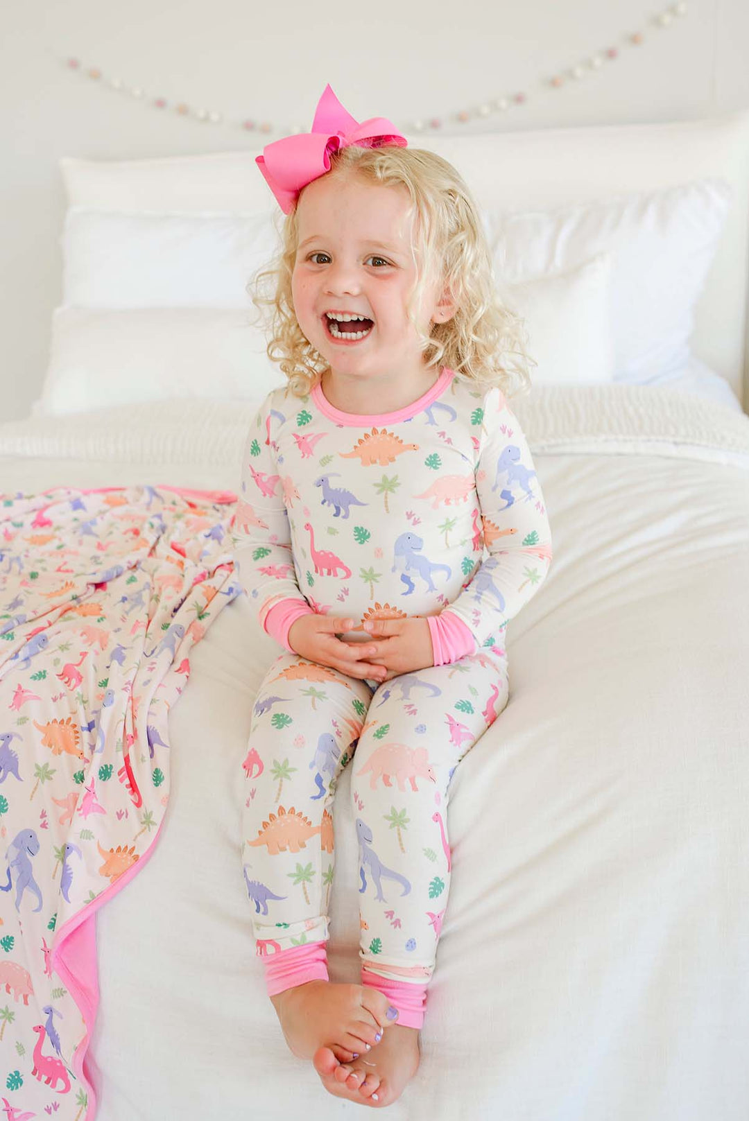 smiling girl sitting on bed in little fossil pink two piece pajama set with the matching bamboo blanket next to her 