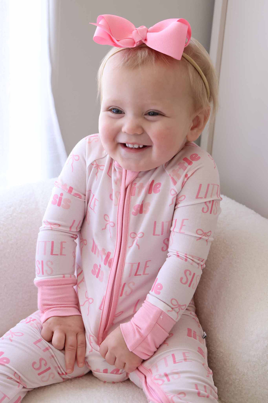 laughing baby girl sitting on fuzzy white chair in little sis convertible zip romper pink with matching ribbon bow headband 
