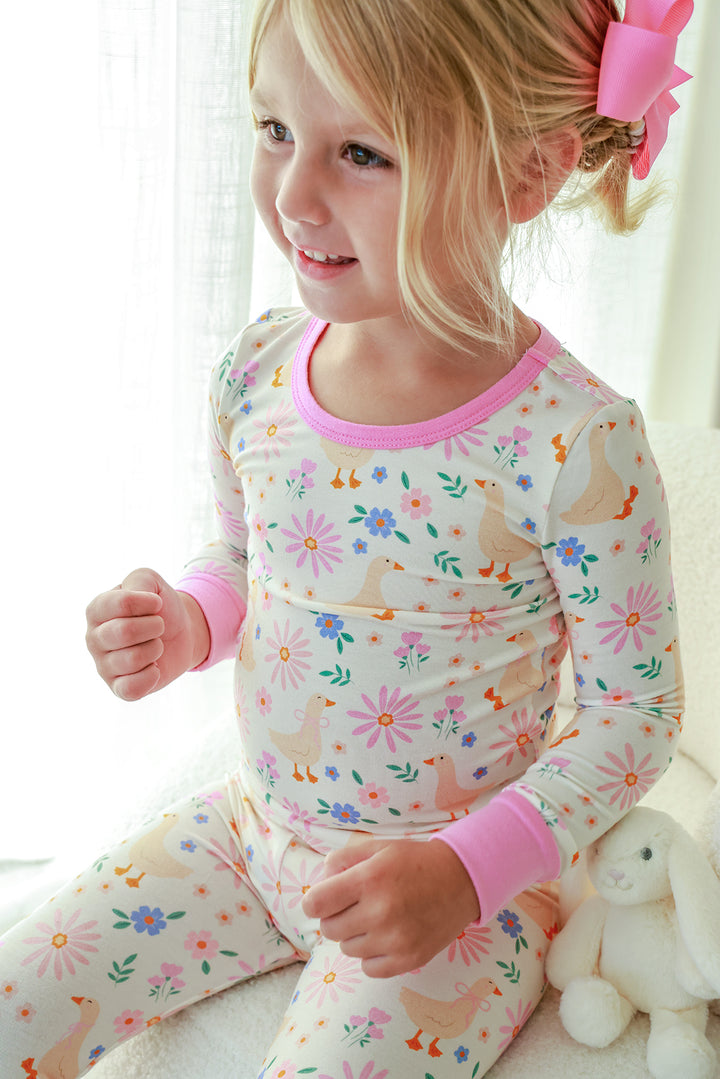 Young girl with pink bow smiling and wearing the Lucy Goosy Two Piece Pajama Set in the floral duck print with pink trim, with a stuffed bunny, lifestyle photo