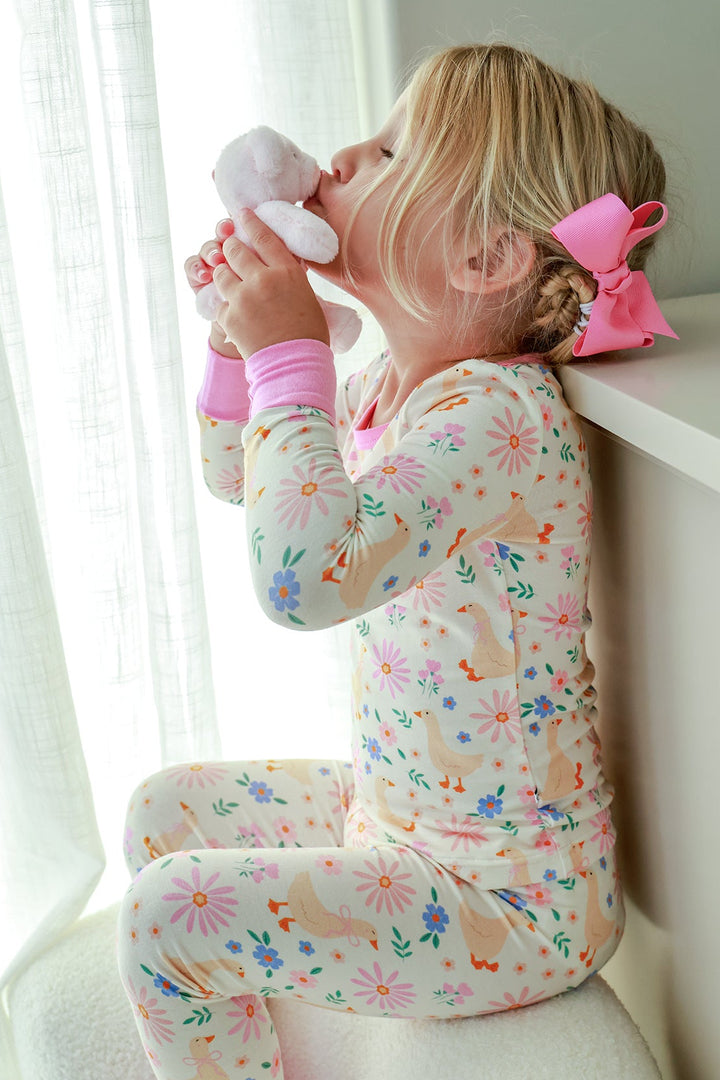 Young girl with pink bow kneeling by a window holding a stuffed animal, wearing the Lucy Goosy Two Piece Pajama Set in the floral duck print, lifestyle photo