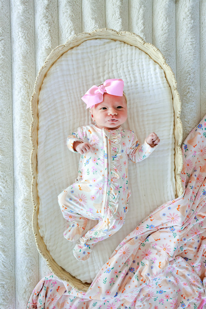 Newborn baby with pink bow lying in a bassinet wearing the Lucy Goosy Ruffle Zipper Footie in the floral duck print, styled with matching floral blanket, lifestyle overhead photo