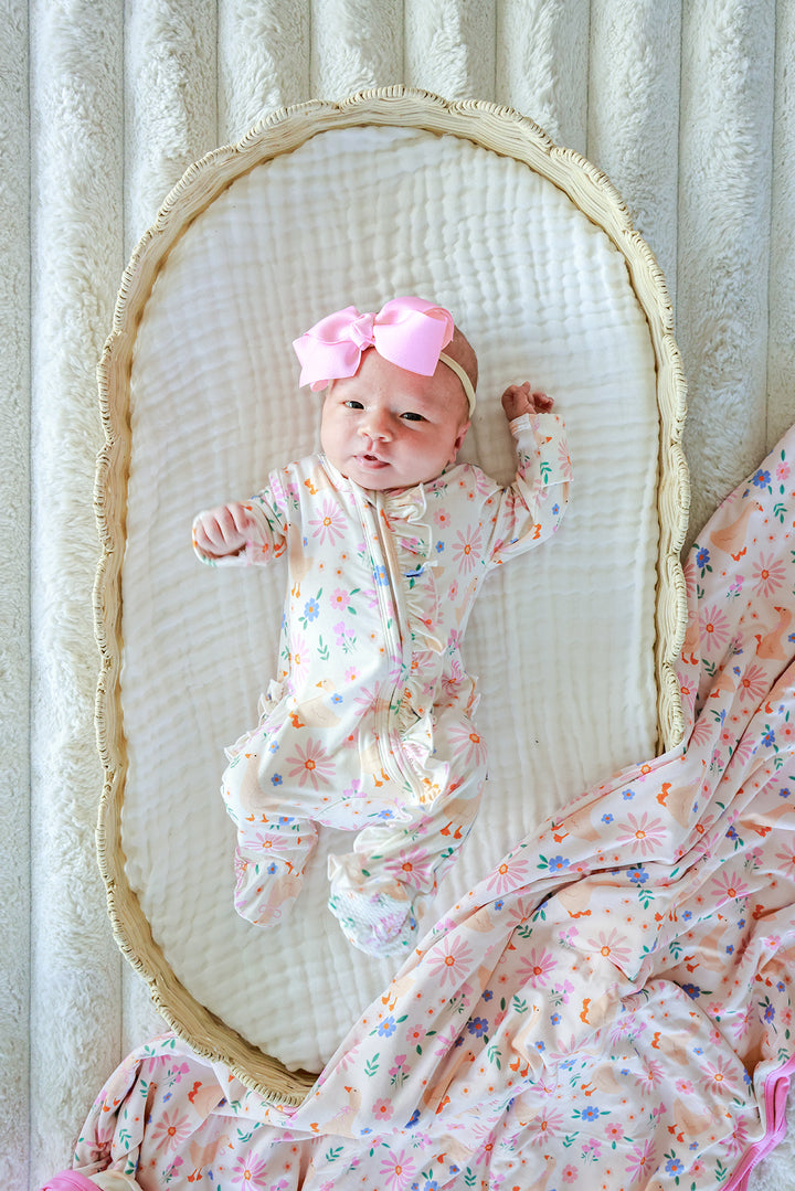 Newborn baby with pink bow lying in a bassinet wearing the Lucy Goosy Ruffle Zipper Footie, styled with a matching floral print blanket, lifestyle photo