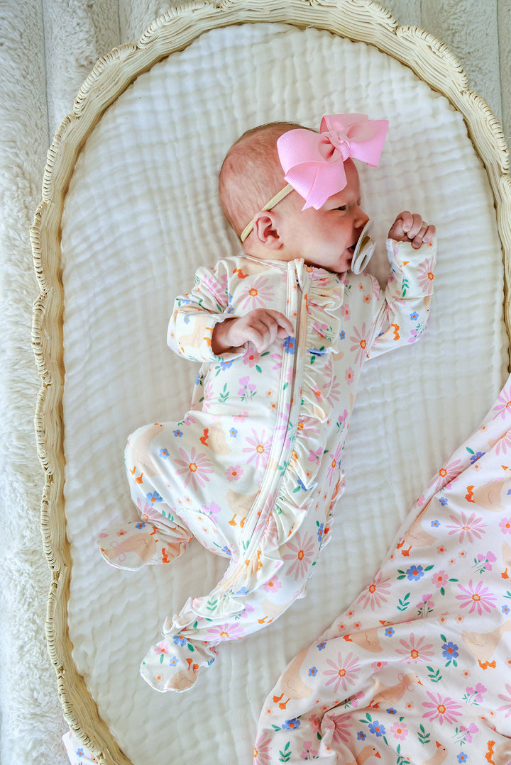 Newborn baby with pink bow lying in a bassinet wearing the Lucy Goosy Ruffle Zipper Footie in the floral duck print, lifestyle overhead photo