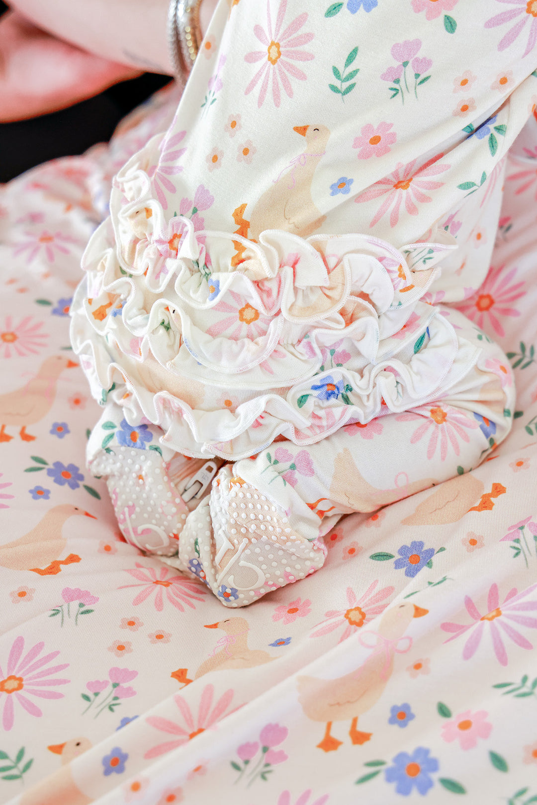 Close-up detail of the Lucy Goosy Ruffle Zipper Footie showing the ruffled waist and decorated footed feet on a matching floral duck print fabric