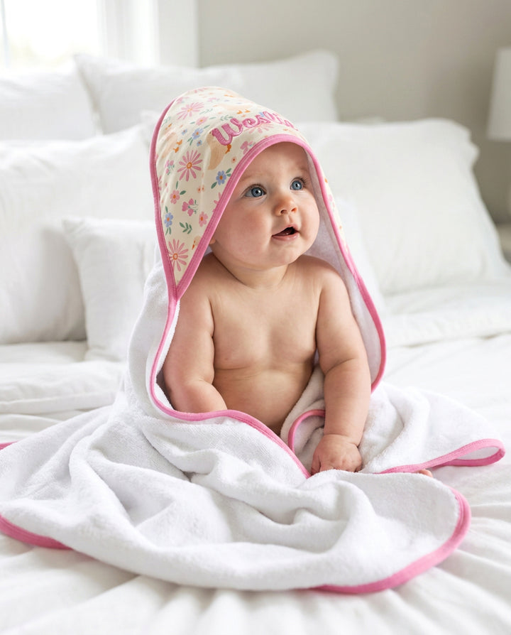 Baby sitting up wearing the Embroidered Bamboo Hooded Towel in the Lucy Goosy floral print with pink trim, displayed on a white bed