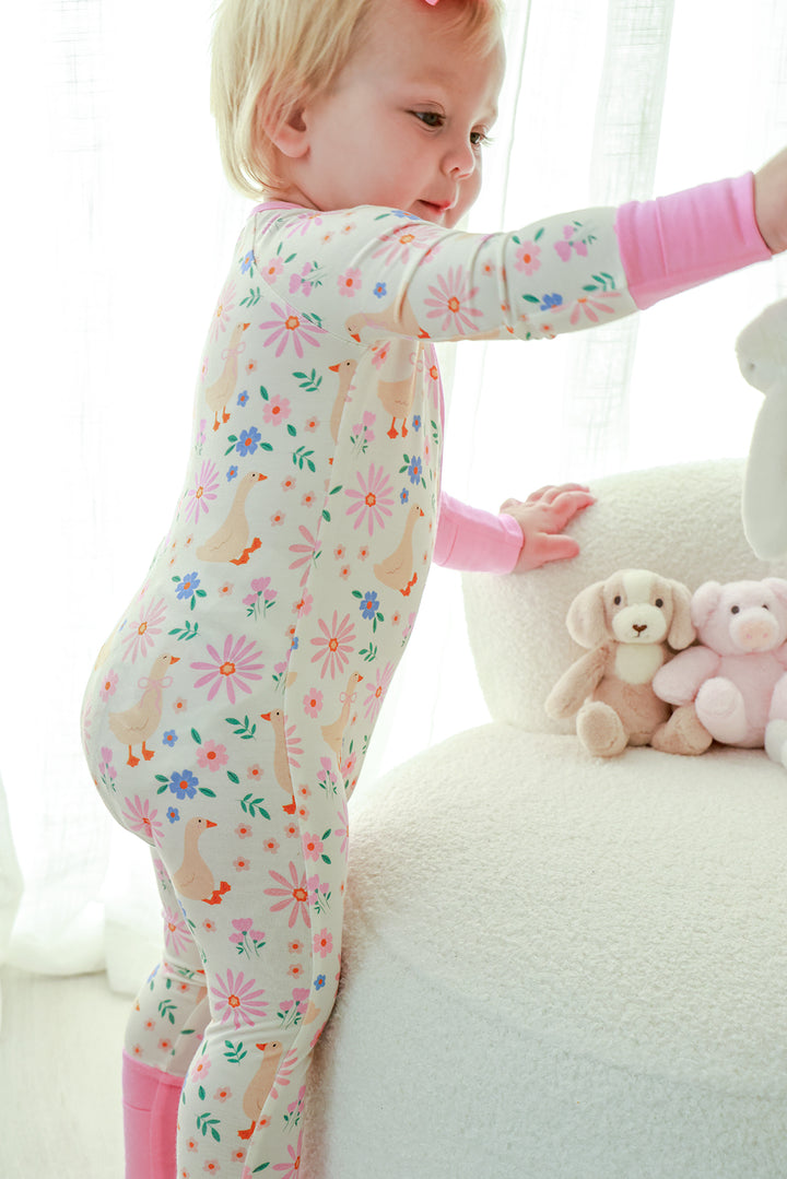 Toddler reaching up while wearing the Lucy Goosy Convertible Zip Romper in the floral duck print with pink cuffs and trim, lifestyle photo with stuffed toys