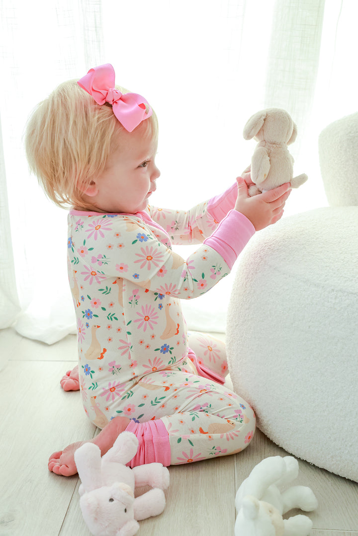 Toddler with pink bow sitting and playing with a stuffed bunny, wearing the Lucy Goosy Convertible Zip Romper in the floral duck print, lifestyle photo