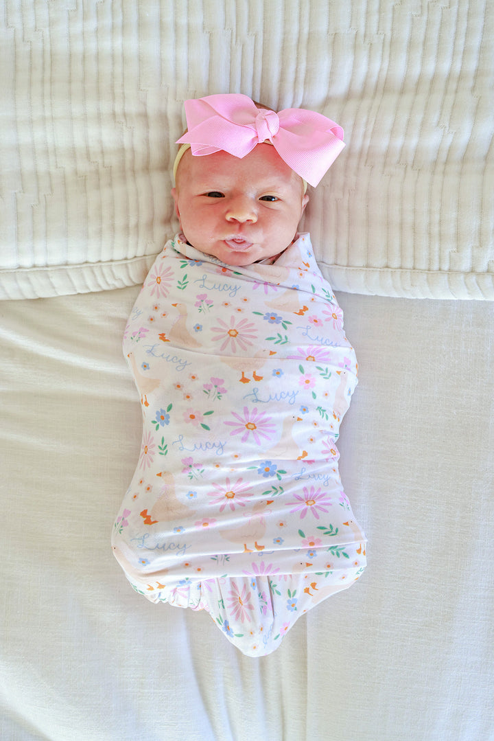 Newborn baby with pink bow swaddled in the Personalized Swaddle Blanket in the Lucy Goosy floral print, lying on a white surface, lifestyle overhead photo