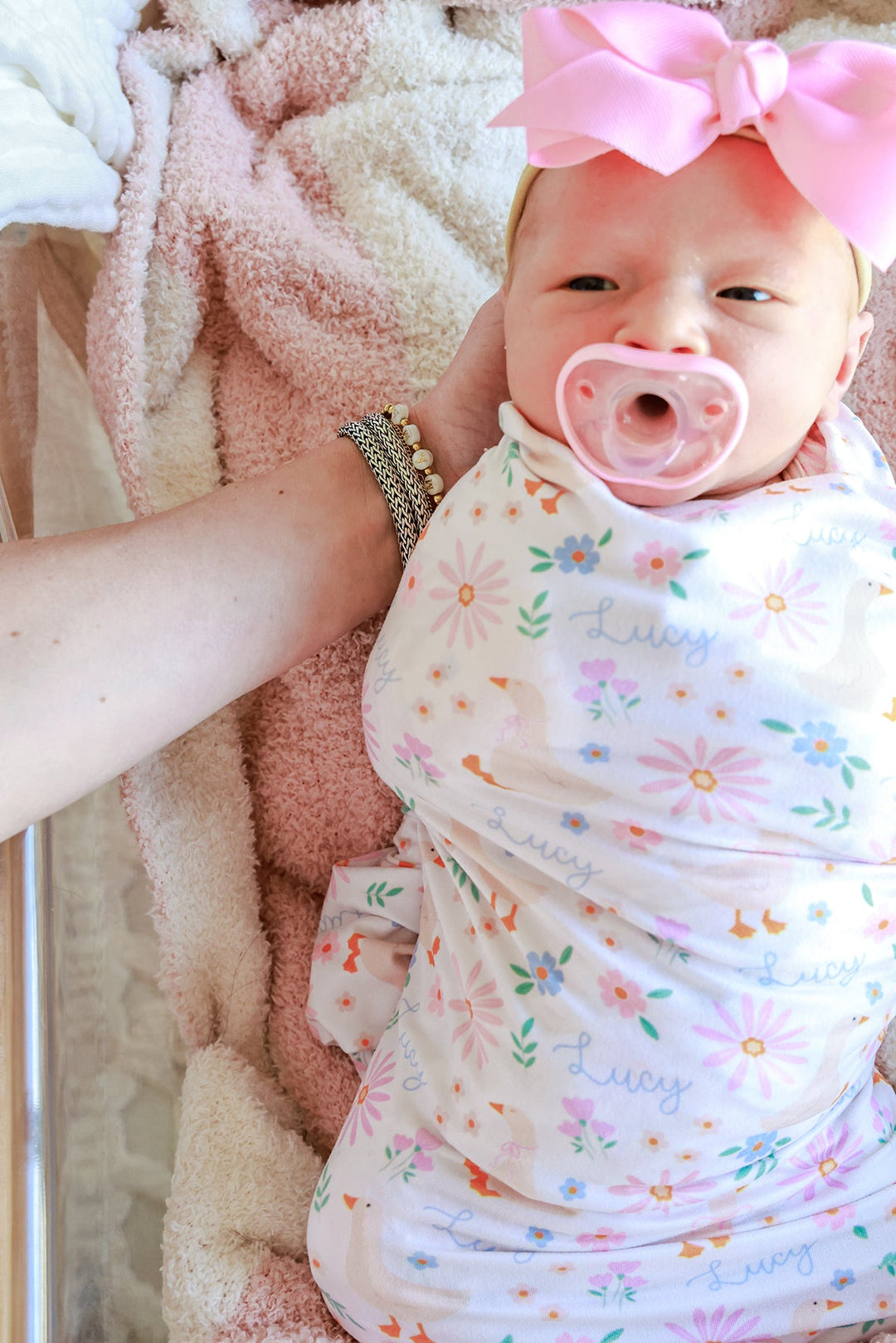 Newborn baby with pink bow and pacifier, swaddled in the Personalized Swaddle Blanket in the Lucy Goosy floral print with name Lucy visible, lifestyle photo