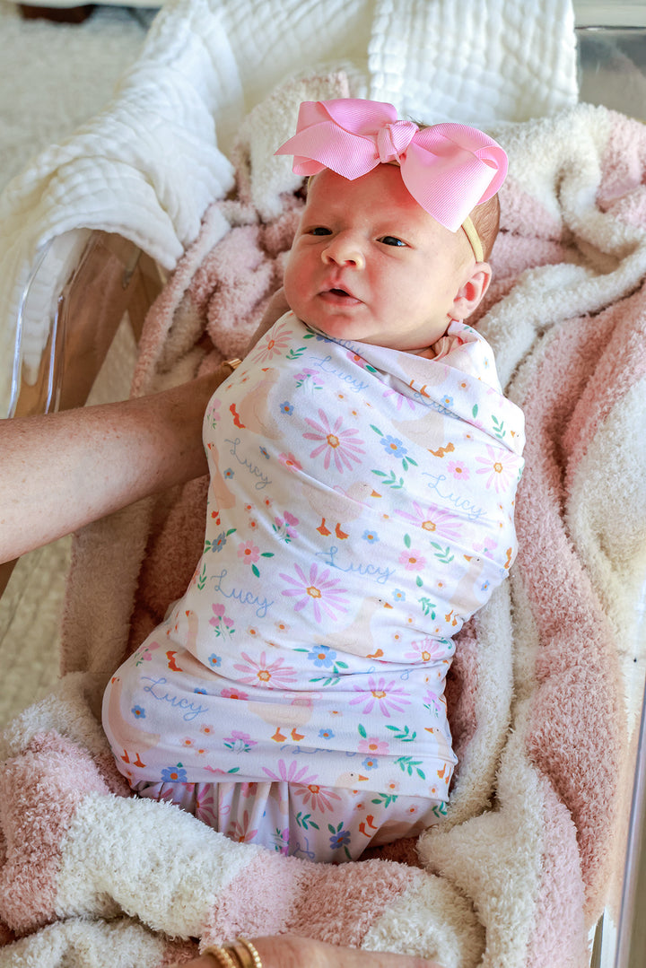 Newborn baby with large pink bow swaddled in the Personalized Swaddle Blanket in the Lucy Goosy floral print, resting on a pink blanket, lifestyle photo