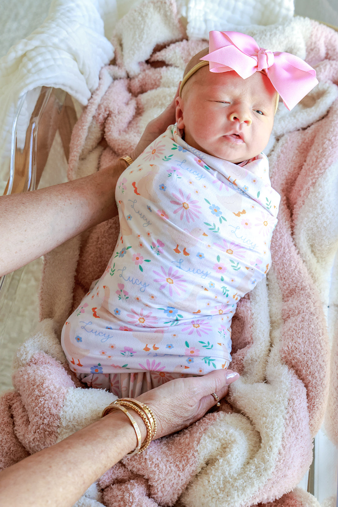 Newborn baby with pink bow swaddled in the Personalized Swaddle Blanket in the Lucy Goosy floral print, held by a parent's hand, lifestyle photo