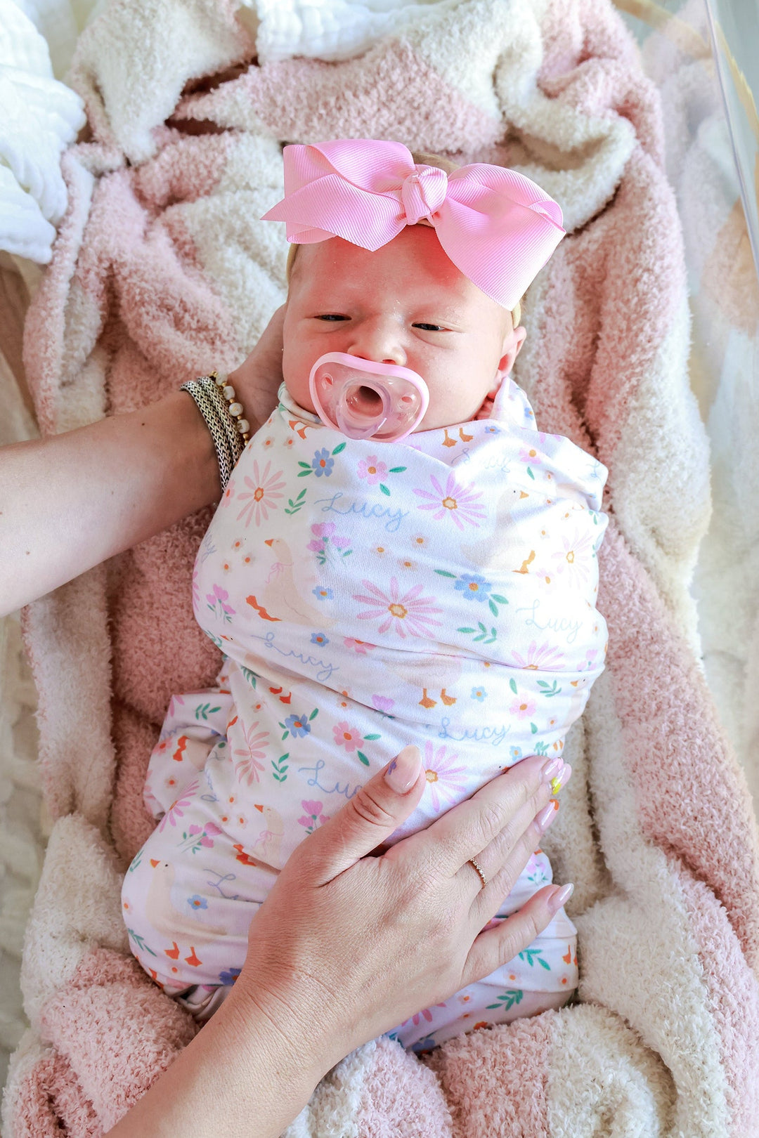 Newborn baby with pink bow swaddled in the Personalized Swaddle Blanket in the Lucy Goosy floral print, being held in a parent's hands, lifestyle photo