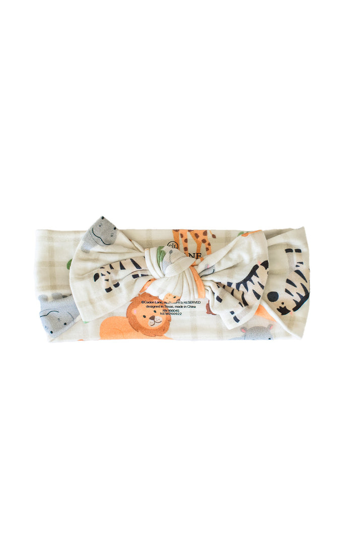 Flat lay of a Caden Lane knotted bow headwrap in a Wild Child print featuring zebras, a lion and hippos on cream plaid.