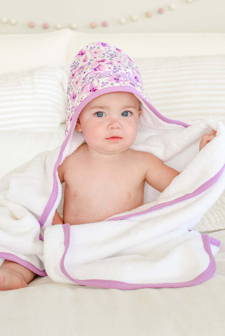 Seated baby gripping the towel edge, wearing a Caden Lane white terry hooded towel in a Lilac Dreams print featuring purple watercolor flowers, reading "Georgia."