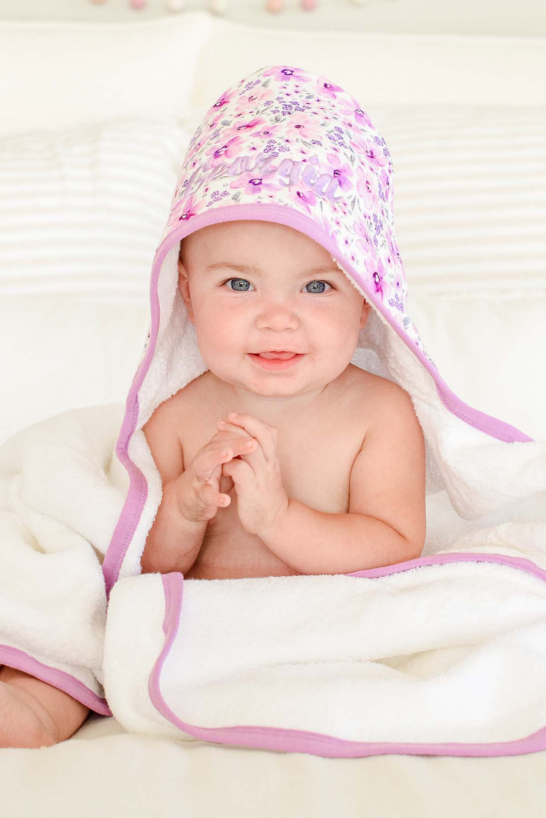 Smiling baby with hands clasped, wearing a Caden Lane white terry hooded towel in a Lilac Dreams print featuring pink watercolor blooms and grey leaves, reading "Georgia."
