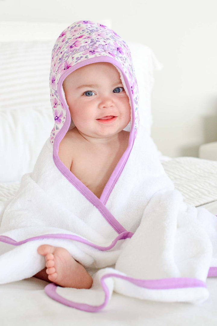 Grinning baby with one bare foot peeking out, wearing a Caden Lane white terry hooded towel with lilac trim in a Lilac Dreams print featuring purple flowers, reading "Georgia."