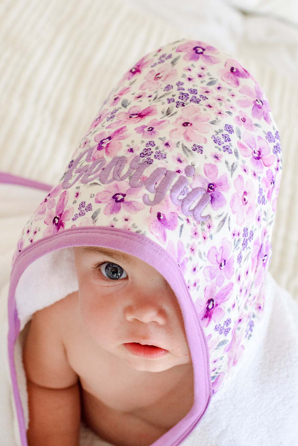 Close-up of a baby peeking out from under the hood of a Caden Lane white terry hooded towel in a Lilac Dreams print featuring purple watercolor flowers, reading "Georgia."