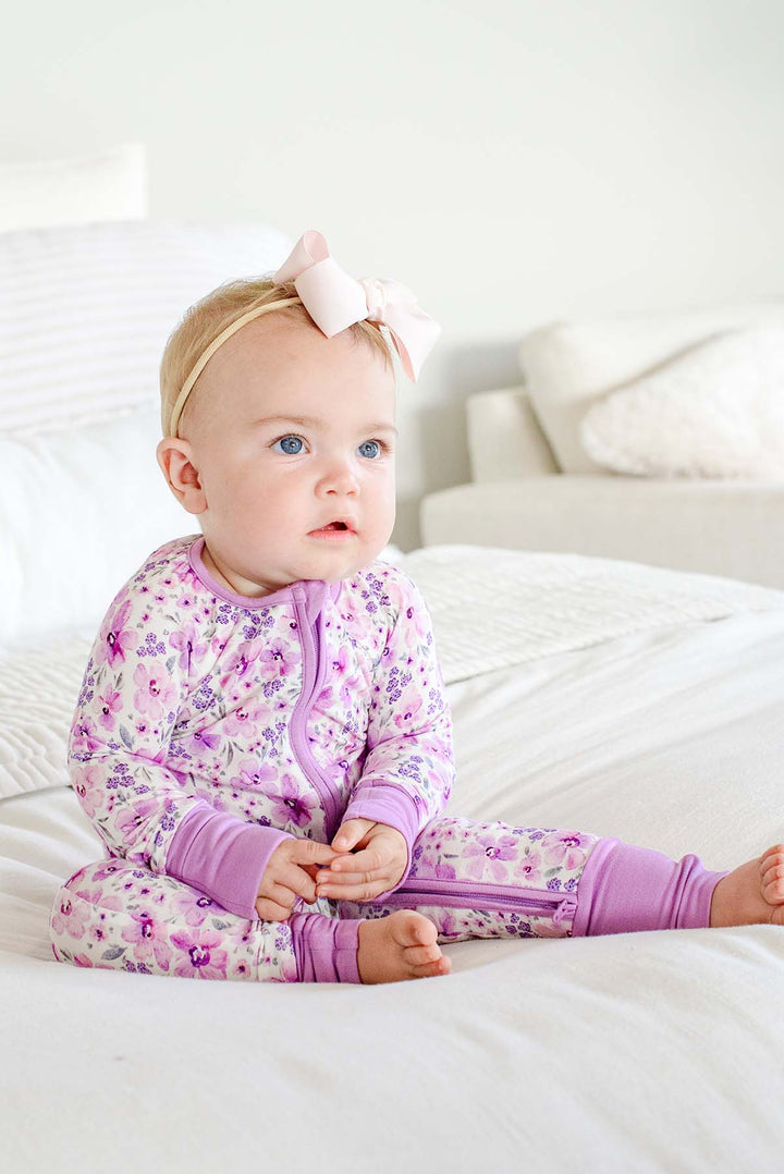 Seated baby with a pale pink bow gazing off camera, wearing a Caden Lane white knit zip romper with lilac cuffs in a Lilac Dreams print featuring purple watercolor flowers.