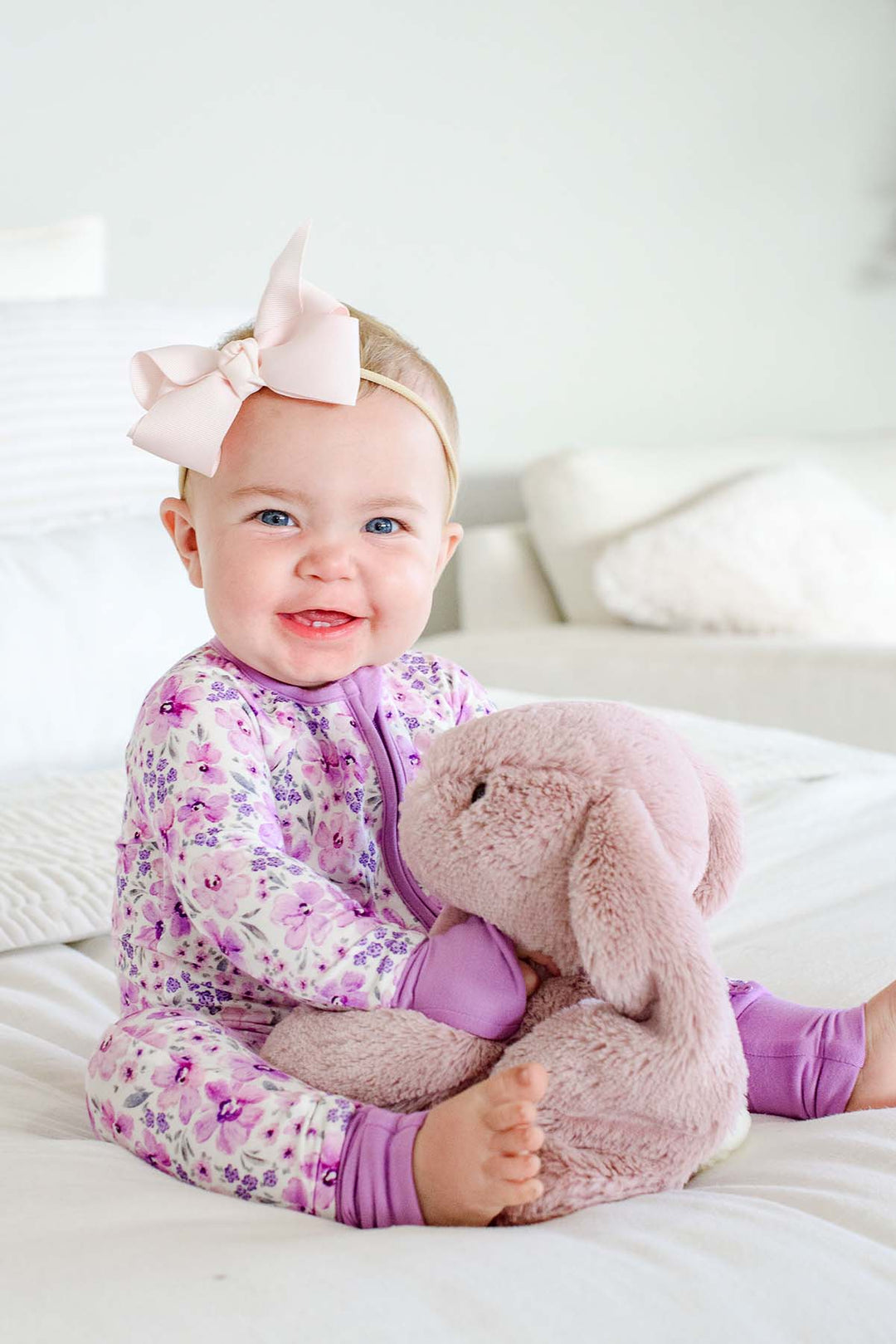 Grinning baby hugging a plush bunny, wearing a Caden Lane white knit zip romper with lilac cuffs in a Lilac Dreams print featuring purple flowers, paired with a pink bow.