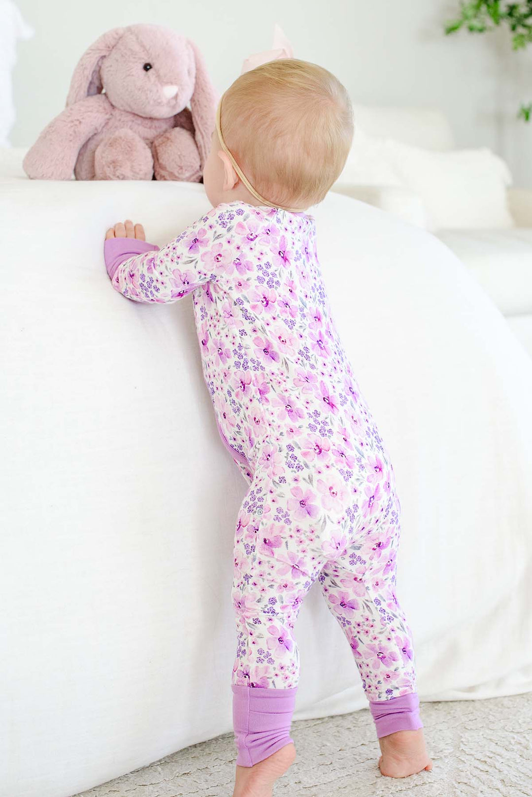 Back view of a standing baby reaching for a plush bunny, wearing a Caden Lane white knit zip romper in a Lilac Dreams print featuring purple flowers.