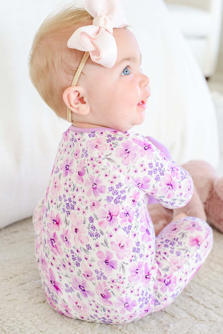 Back view of a seated baby looking up over her shoulder, wearing a Caden Lane white knit zip romper in a Lilac Dreams print featuring pink watercolor blooms.