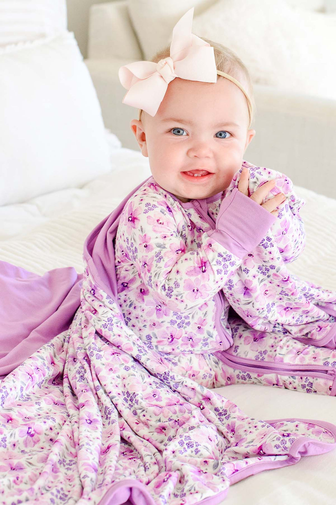 A grinning baby clutching a corner of a Caden Lane cream knit blanket with a lilac reverse in a Lilac Dreams print, paired with a matching zip romper.