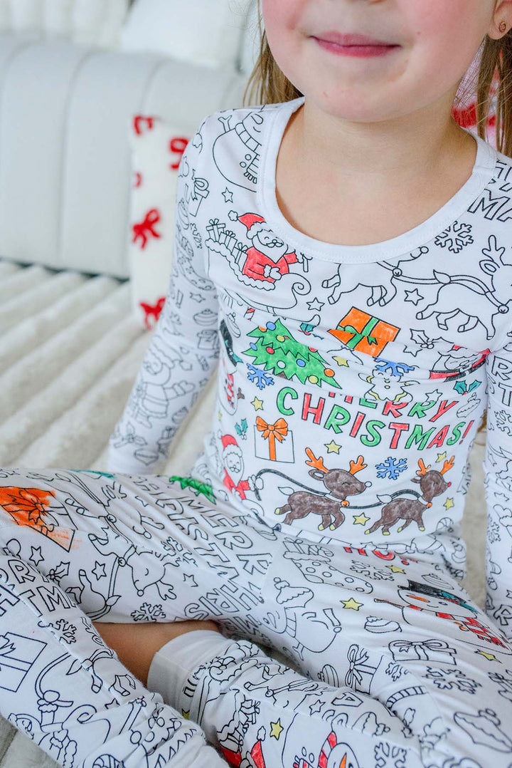 Close-up of a young girl wearing Caden Lane's Merry Christmas color-in pajama set, featuring a black-and-white line art design with Santa, reindeer, and "Merry Christmas" text partially colored in with red and green marker.