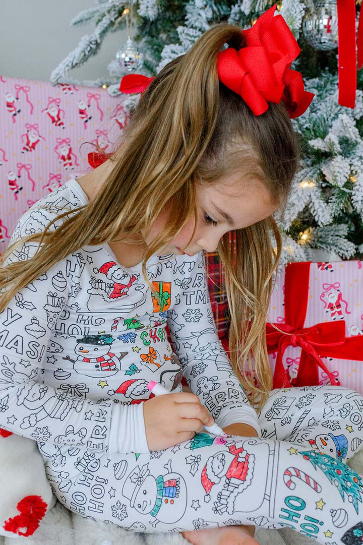 Girl sitting cross-legged in Caden Lane color-in Christmas pajamas, using a fabric marker to color a snowman design on the leggings, with a Caden Lane fabric marker set and Christmas gifts in the background