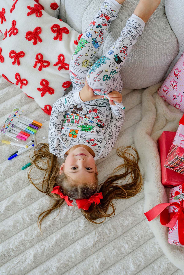 Overhead shot of a girl lying on a rug in Caden Lane color-in Christmas pajamas, holding up her leg to color a "Ho Ho Ho" leggings design, red bows in her pigtails.