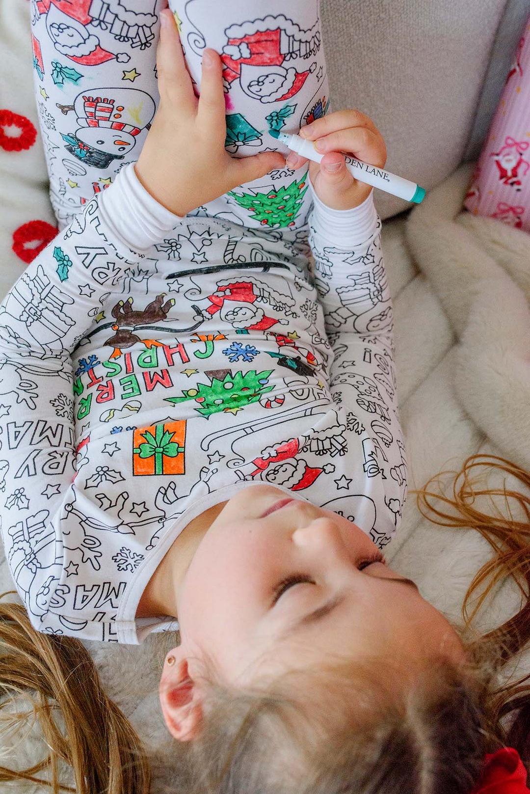 Close-up overhead view of a girl coloring the "Merry Christmas" text and holiday graphics on her Caden Lane pajama top with a teal marker.