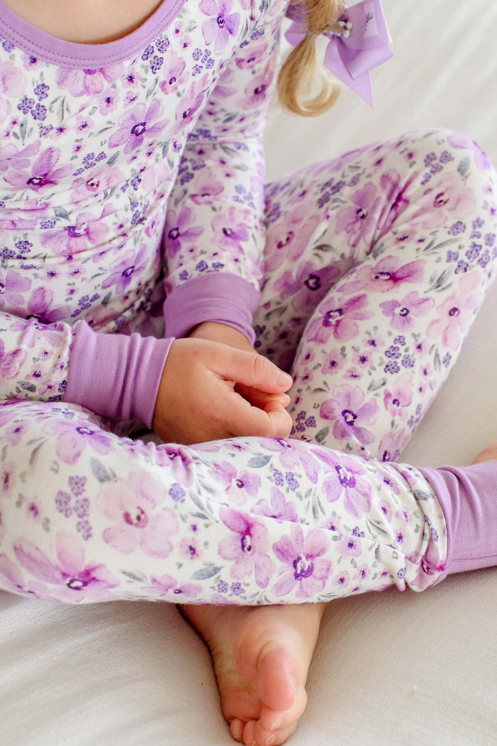 Close-up of a girl sitting cross-legged in Caden Lane cream knit pajamas in a Lilac Dreams print featuring watercolor flowers, with lilac cuffs.